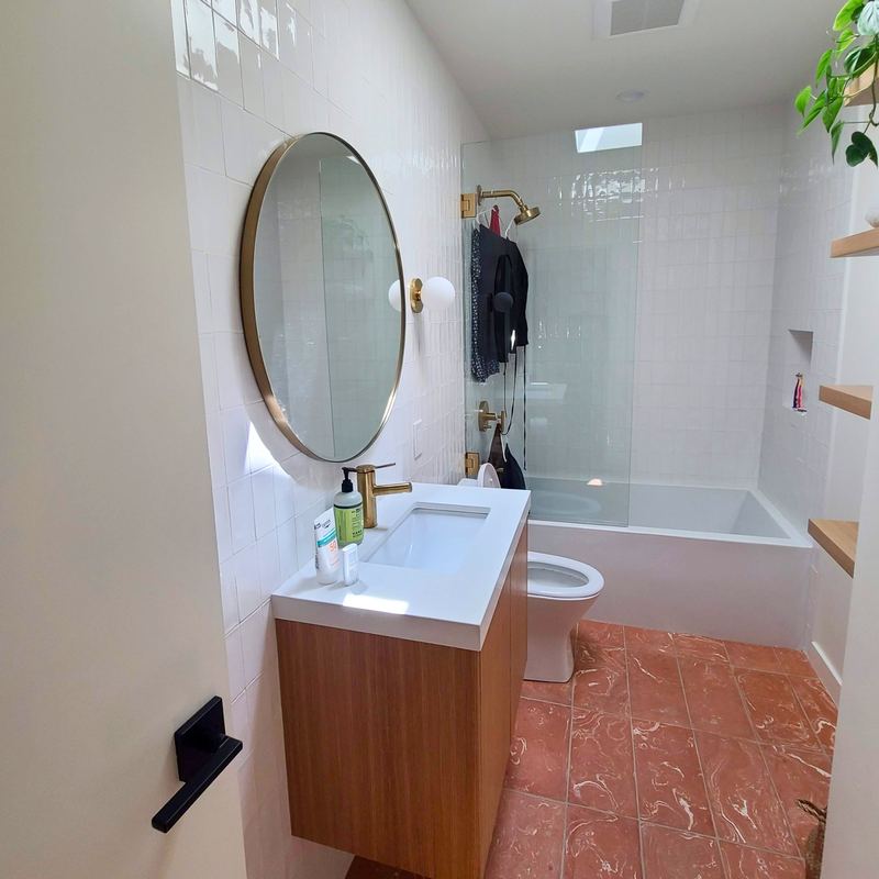 Guest bathroom with white stacked tile, soaking tub, glass panel, terracotta floor and round brass mirror